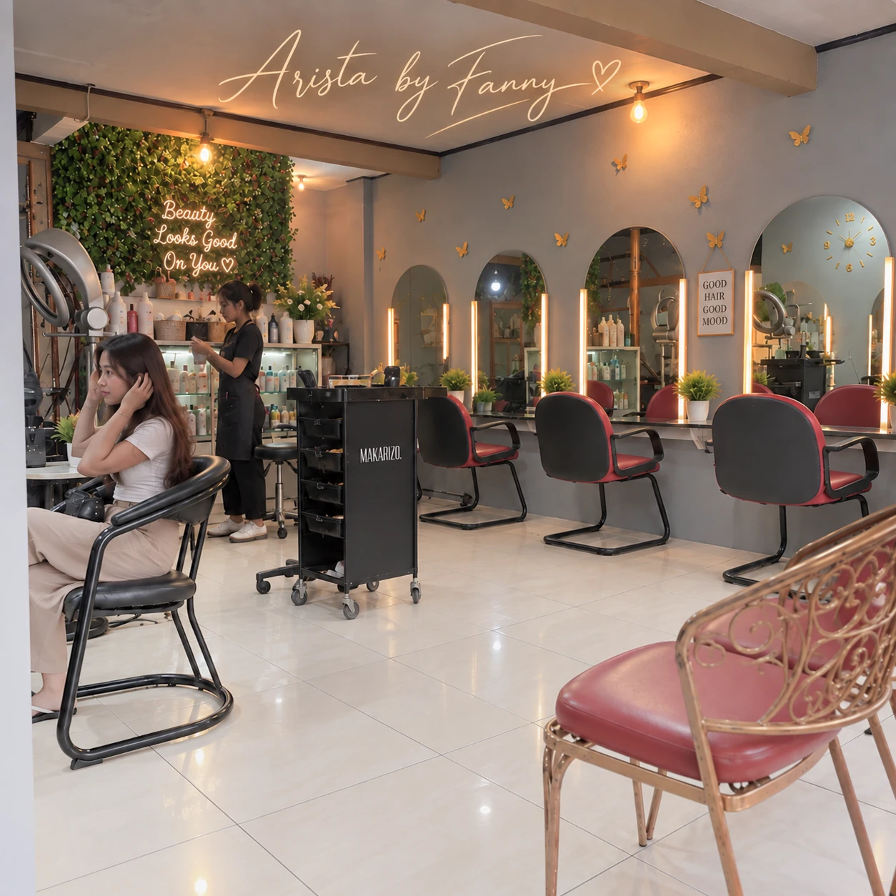 Ruang dalam Arsita Salon di Pemalang: dinding tanaman hijau dengan tulisan neon, cermin lengkung berlampu, kursi salon merah, dan seorang pelanggan yang sedang dilayani.